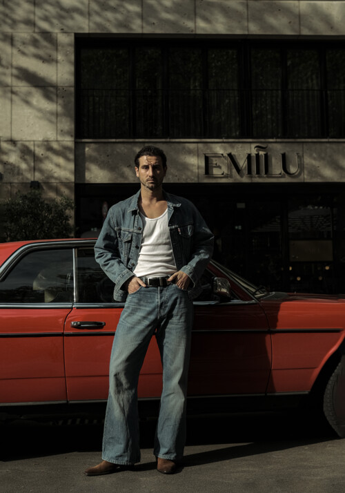 Mann im Jeans look lehnt an ein rotes Auto vor einem Geb&auml;ude mit der Aufschrift EmiLu